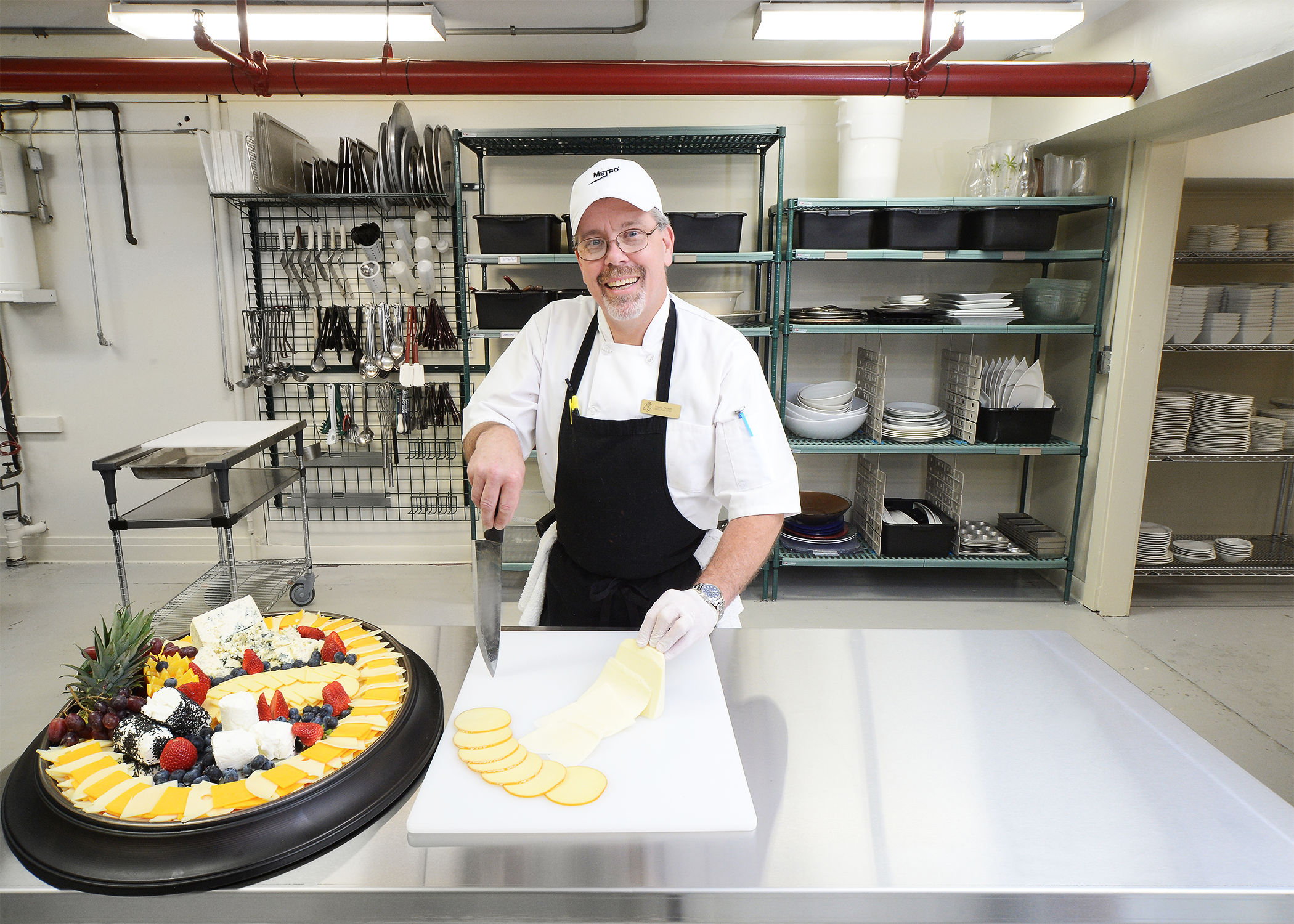 A knife wielding man wearing an apron smiles towards us while standing behind a table holding a party tray and cutting tray of cheese.