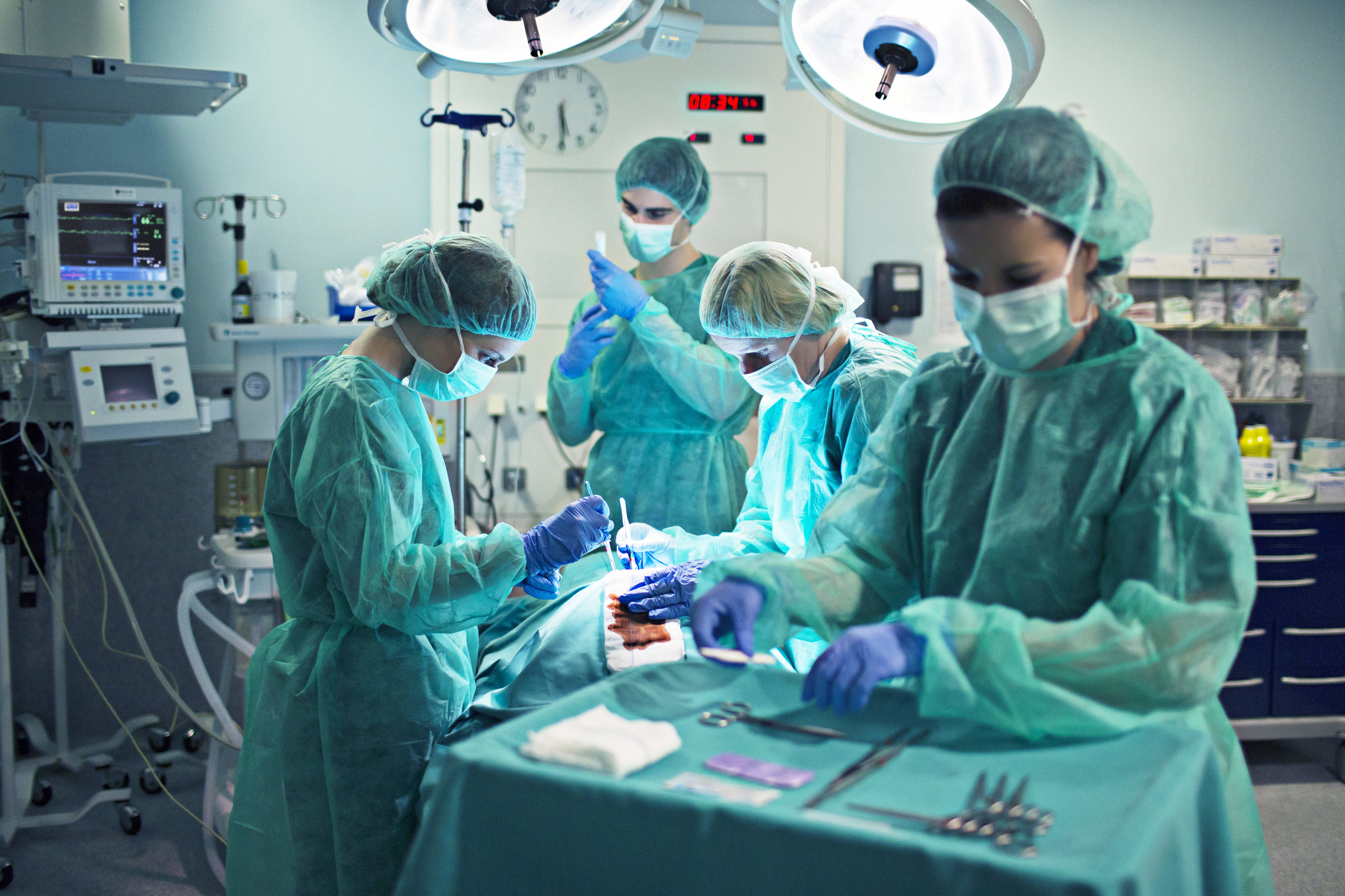 In the background: three nurses hover around a patient while holding medical equipment. Foreground: a nurse is selecting medical equipment organized atop a procedure cart .